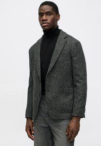 Hallhalli herringbone mustriga bleiser, mis on valmistatud tekstureeritud kangast, sellel on klapiga krae, üks nööbiga kinnitamine ja kaks esist pocketit, kantud musta kõrge kaelusega tossuga.