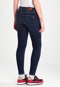 Jeans skinny azul oscuro con una textura de denim suave, que cuentan con cinco bolsillos y una etiqueta de cuero marrón claro en la cintura. Llevados con zapatillas rosas.
