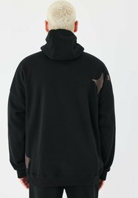 Sort oversized hoodie med en dobbeltlaget hætte, glat tekstur og kontrasterende brune accenter på skuldrene. Slanke ribbede manchetter.