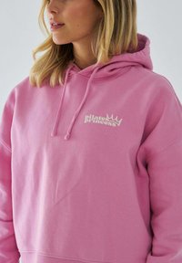 Różowy sweatshirt z kapturem, posiadający sznurki i grafikę na przodzie "Pilates Princess." Wykonany z miękkiego materiału o luźnym kroju.