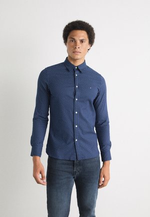 Camisa de manga larga azul marino con un pequeño patrón de lunares blancos, cuello abotonado y tapeta delantera. Combinada con unos vaqueros oscuros.