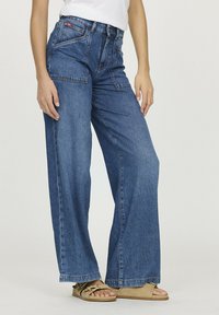 Jean en denim bleu à taille moyenne et jambes larges, avec cinq poches et une fermeture à bouton. Le tissu a une texture lisse et présente des décolorations légères.