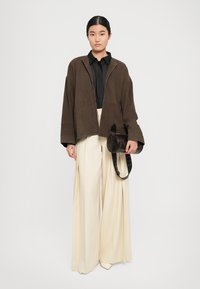 Femme portant une veste marron ample, un shirt noir, un pantalon large crème, tenant un sac cylindrique marron foncé, debout devant un fond uni.
