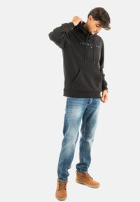 Felpa nera con cappuccio e lacci, tasca frontale, testo del logo sul petto, abbinata a jeans blu medio e stivali beige. Design semplice e casual.