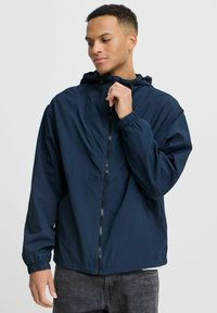 Veste coupe-vent navy avec capuche, poignets élastiques et fermeture éclair sur le devant. Fabriquée en tissu léger et lisse, adaptée pour les vêtements d'extérieur.