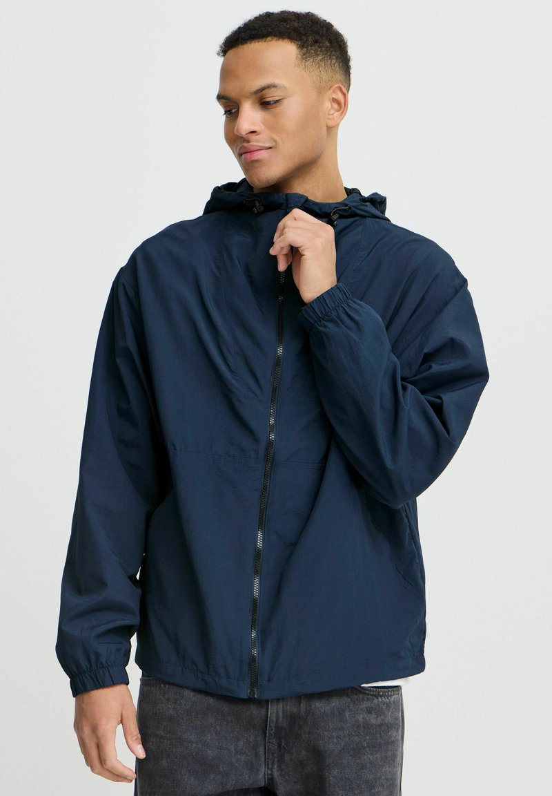 Veste coupe-vent navy avec capuche, poignets élastiques et fermeture éclair sur le devant. Fabriquée en tissu léger et lisse, adaptée pour les vêtements d'extérieur.