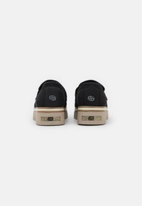 Black slip-on shoes with a padded collar, tan rubber sole, and a logo on the back. Minimalistic design, sturdy material, and smooth texture.