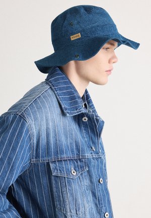 Blauer Denim-Bucket Hat mit breiter Krempe, versehen mit einem Logo-Patch und drei Druckknöpfen; getragen mit einer passenden gestreiften Denim-Jacke.