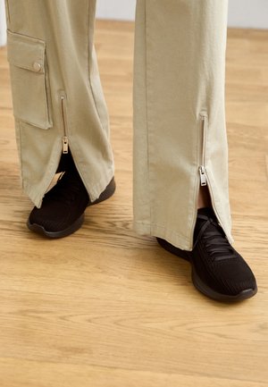 Pantalones beige de pierna ancha con cremalleras en los tobillos, combinados con zapatillas deportivas negras con parte superior texturizada y suela acolchada.