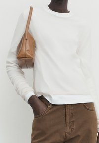 Vit crewneck tröja i mjukt tyg, matchad med bruna byxor. En liten brun handväska hålls under armen.