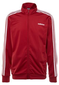 Chaqueta deportiva roja con cremallera hecha de tejido suave, presenta acentos en blanco con tres rayas a lo largo de las mangas y un logo de marca en el lado izquierdo del pecho.