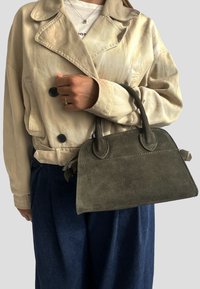 Olivově zelená semišová kabelka se strukturovaným tvarem a dvěma krátkými uchy, kterou drží osoba v béžovém saku a modrých kalhotách.
