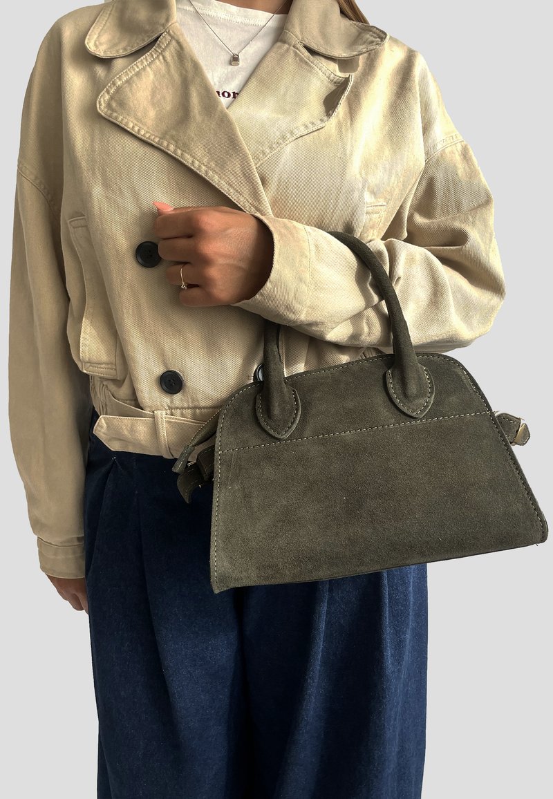 Olivově zelená semišová kabelka se strukturovaným tvarem a dvěma krátkými uchy, kterou drží osoba v béžovém saku a modrých kalhotách.