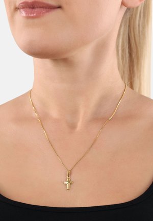 Gouden kruis hangerketting op een dunne ketting, met een gepolijste afwerking en verfijnde detailing, gedragen tegen blote huid met een donker topje.