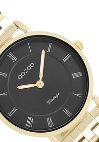 Montre-bracelet dorée avec un cadran noir, des chiffres romains et deux aiguilles dorées. Bracelet en style maillon avec une finition polie.