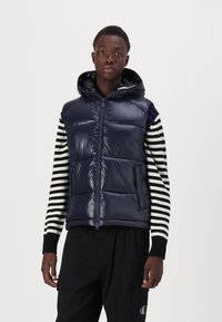 Vesta puffer navy cu glugă, cu un aspect lucios, cu închidere prin fermoar și o buzunar. Pur și simplu peste un tricou cu mânecă lungă în dungi alb-negru.