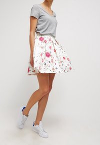 Blumendruckrock in Weiß mit pinken und grünen Akzenten, aus leichtem Stoff; kombiniert mit einem grauen V-Ausschnitt-T-Shirt und weißen Sneakers.
