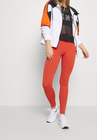 Completo sportivo con una maglietta nera in rete con lettering bianco, una giacca arancione e bianca con zip, e leggings rossi aderenti con scarpe da ginnastica bianche.