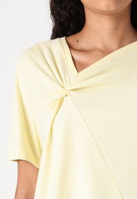 Žlutý top s krátkým rukávem, který má drapovaný design a zkroucený detail u výstřihu, vyrobený z měkké tkaniny pro pohodlí.