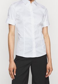 Chemise blanche à manches courtes avec boutons et col, présentant un design élégant, une texture lisse et une coupe ajustée. Associée à un pantalon noir.