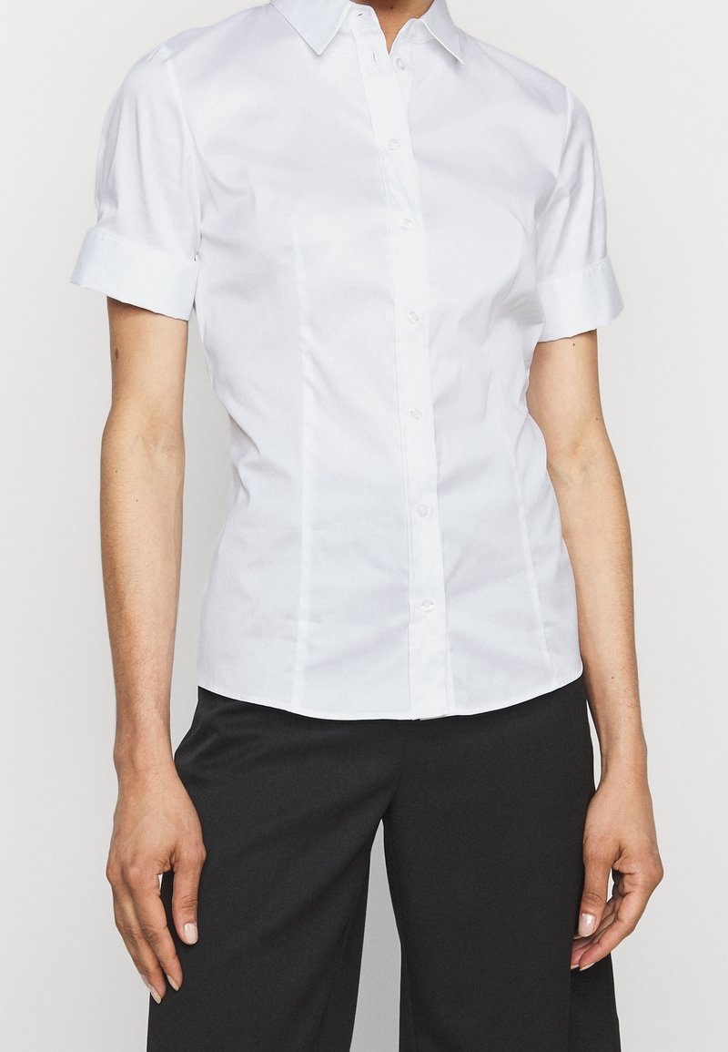Chemise blanche à manches courtes avec boutons et col, présentant un design élégant, une texture lisse et une coupe ajustée. Associée à un pantalon noir.