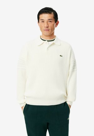 Mann trägt ein weißes, geripptes langärmeliges Poloshirt und dunkle Hose, die Hände in den Taschen, steht vor einem einfarbigen hellen Hintergrund.