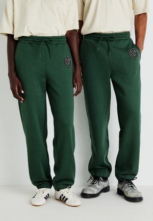 Los pantalones de chándal verdes presentan una cintura elástica con cordón, puños acanalados y un parche de logo circular en la parte frontal. Textura de tela suave.