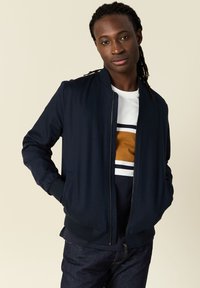 Marinblå bomberjacka med frontdragkedja, ribbade muddar och nederkant. Bärs över en vit skjorta med brun och marinblå horisontella ränder.