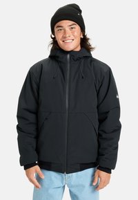 Jeune homme souriant, portant un bonnet noir, une veste à capuche noire avec fermeture éclair et un jean bleu clair, se tenant devant un fond blanc.