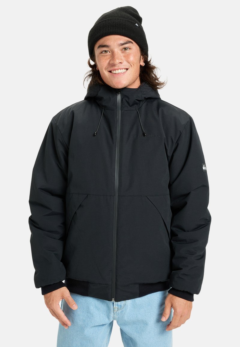 Jeune homme souriant, portant un bonnet noir, une veste à capuche noire avec fermeture éclair et un jean bleu clair, se tenant devant un fond blanc.