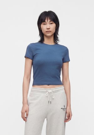 Giovane donna con capelli neri fino alle spalle indossa una t-shirt blu aderente corta e pantaloni della tuta grigio chiaro con coulisse su sfondo bianco.