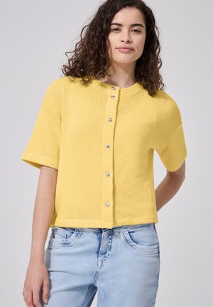 Frau mit lockigem dunklem Haar, die einen gelben kurzärmeligen, geknöpften Pullover und hellblaue Jeans vor einem schlichten Hintergrund trägt.