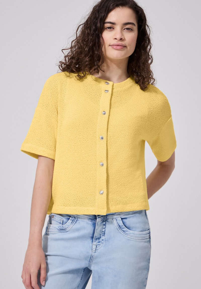 Femme aux cheveux bouclés foncés portant un pull jaune à manches courtes boutonné et un jean bleu clair sur un fond uni.