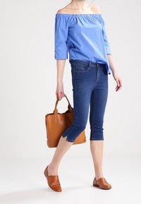 Blusa blu off-the-shoulder con maniche a tre quarti, abbinata a jeans scuri a vita alta. Borsa a tracolla marrone e scarpe slip-on marroni completano il look.