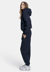 Nuori nainen tummansinisessä hupparissa ja verryttelyhousuissa, käsi taskussa, seisoo sivuttain valkoiset tennarit jalassa yksinkertaisella taustalla.