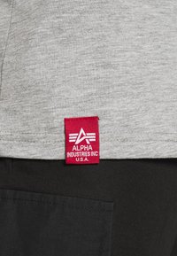 Szürke pólóanyag, piros címkével, amelyen az "ALPHA INDUSTRIES INC." felirat és egy logó látható, az oldalára varrva. Lent egy fekete zseb látható.