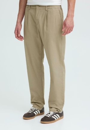 Person trägt beige Freizeithose mit Knopf und elastischem Bund, kombiniert mit schwarzen und weißen Turnschuhen.