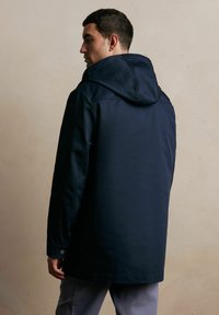 Granatowa kurtka wodoodporna z kapturem, posiadająca długie rękawy, wysoki kołnierz oraz ozdobne guziki na mankietach, teksturowany materiał oraz średnią długość.