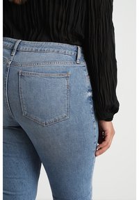 Femme portant un jean en denim bleu clair avec une poche arrière, associée à un chemisier noir texturé à manches longues, la main reposant le long du corps.