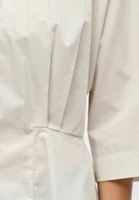 Gros plan sur un vêtement beige montrant un détail de tissu froncé et une manche courte sur un bras.