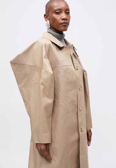 Béžový trench coat s oversized, strukturovanými rukávy, se zlatými knoflíky a klasickým límcem. Hladká textilie s viditelnými švy.