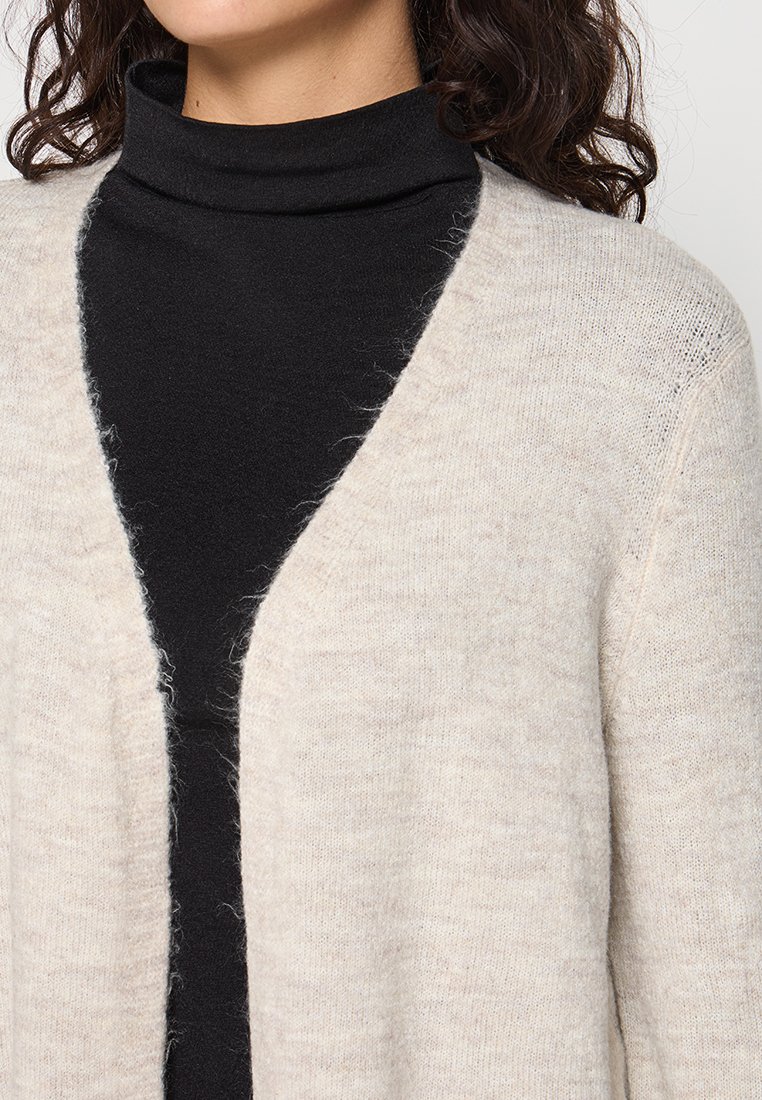 Beige gebreide cardigan met een zachte, pluizige textuur, met een open voorkant en drop shoulders, gelaagd over een zwarte coltrui.