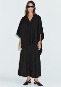 Femme portant un poncho noir à col en V, une jupe longue plissée, des chaussures noires à petit talon, des lunettes de soleil foncées et un bracelet en argent sur un fond uni.