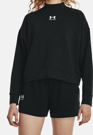 Kvinde iført sort Under Armour langærmet sweatshirt og matchende shorts, stående mod en ensartet lys baggrund.