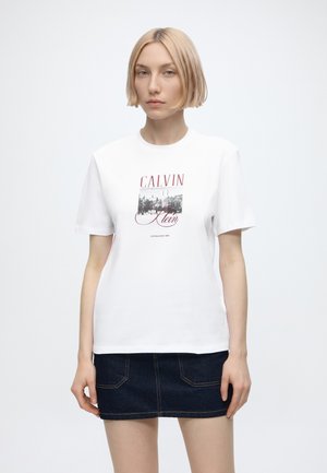 Bleg kvinde iført en hvid Calvin Klein T-shirt med rød tekst og et sort-hvidt motiv, kombineret med en mørk denim mini nederdel.
