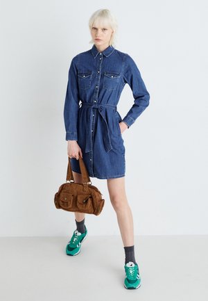 Denim shirt dress with long sleeves, button closure, two chest pockets, a tied waist, and a brown fabric handbag. Model wears green sneakers.