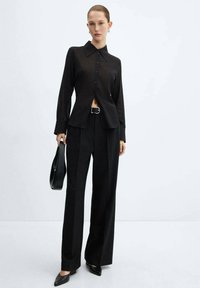 Vrouw in zwarte blouse met knopen, wijde broek en een zwarte handtas, staande tegen een effen lichte achtergrond.