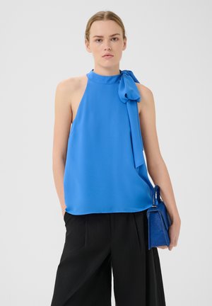 Blauer Trägeroberteil aus weichem Stoff mit einer Bindungsdetail am Hals, kombiniert mit lockeren schwarzen Hosen und einer kleinen blauen Handtasche.
