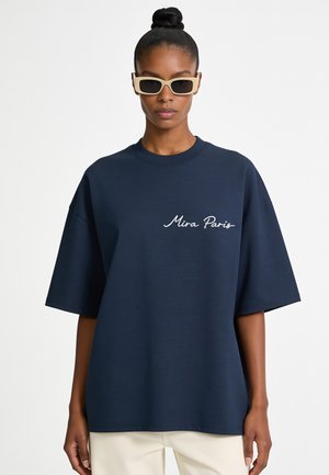 Vrouw met een donkere huid en haar in een knot draagt beige zonnebril, een navyblauw oversized t-shirt met tekst "Mira Paris" en lichte broek.