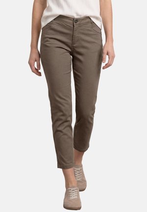 Kobieta w brązowych skróconych spodniach i beżowych trampkach, stojąca na białym tle.